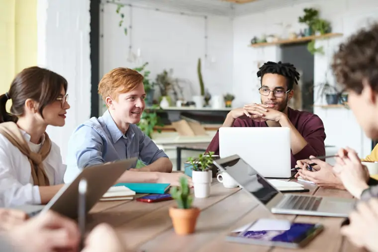 Buona comunicazione in azienda tra più collaboratori seduti allo stesso tavolo che dialogano di fronte ai loro laptop | MentiPratiche